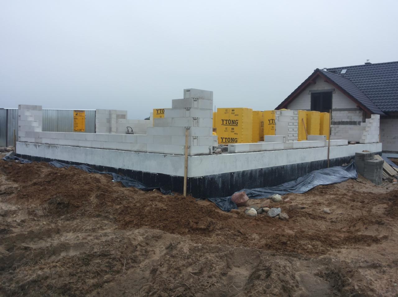 Wznoszenie ścian parteru domu z bloczków Ytong na fundamencie z hydroizolacją, widoczne palety z bloczkami i fragment dachu sąsiedniego budynku w pochmurny dzień.