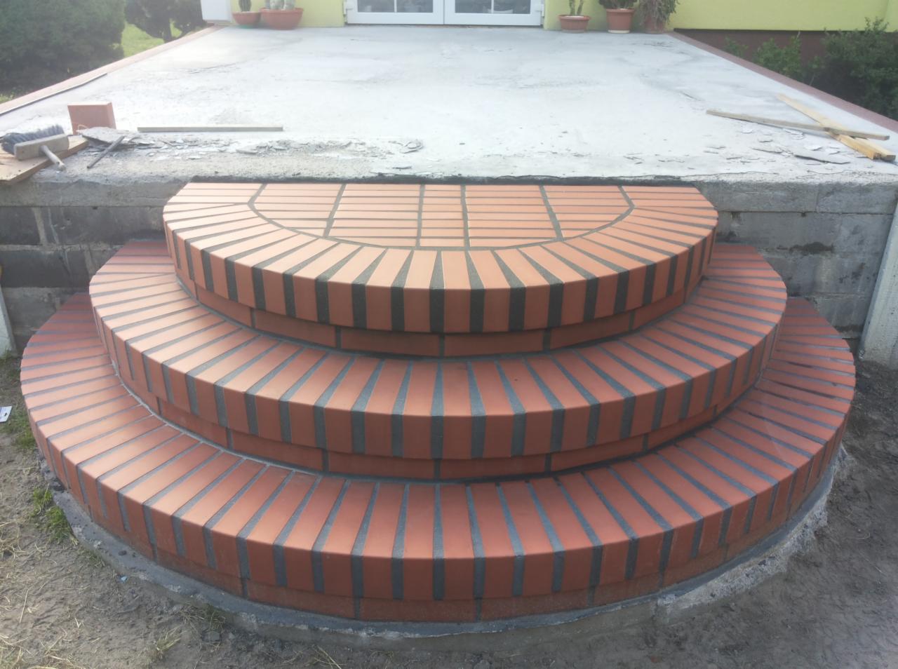 Półokrągłe, trójstopniowe schody wejściowe wyłożone czerwoną cegłą klinkierową z ciemnymi fugami, prowadzące do betonowego podestu przed budynkiem z białymi drzwiami i roślinami w donicach.