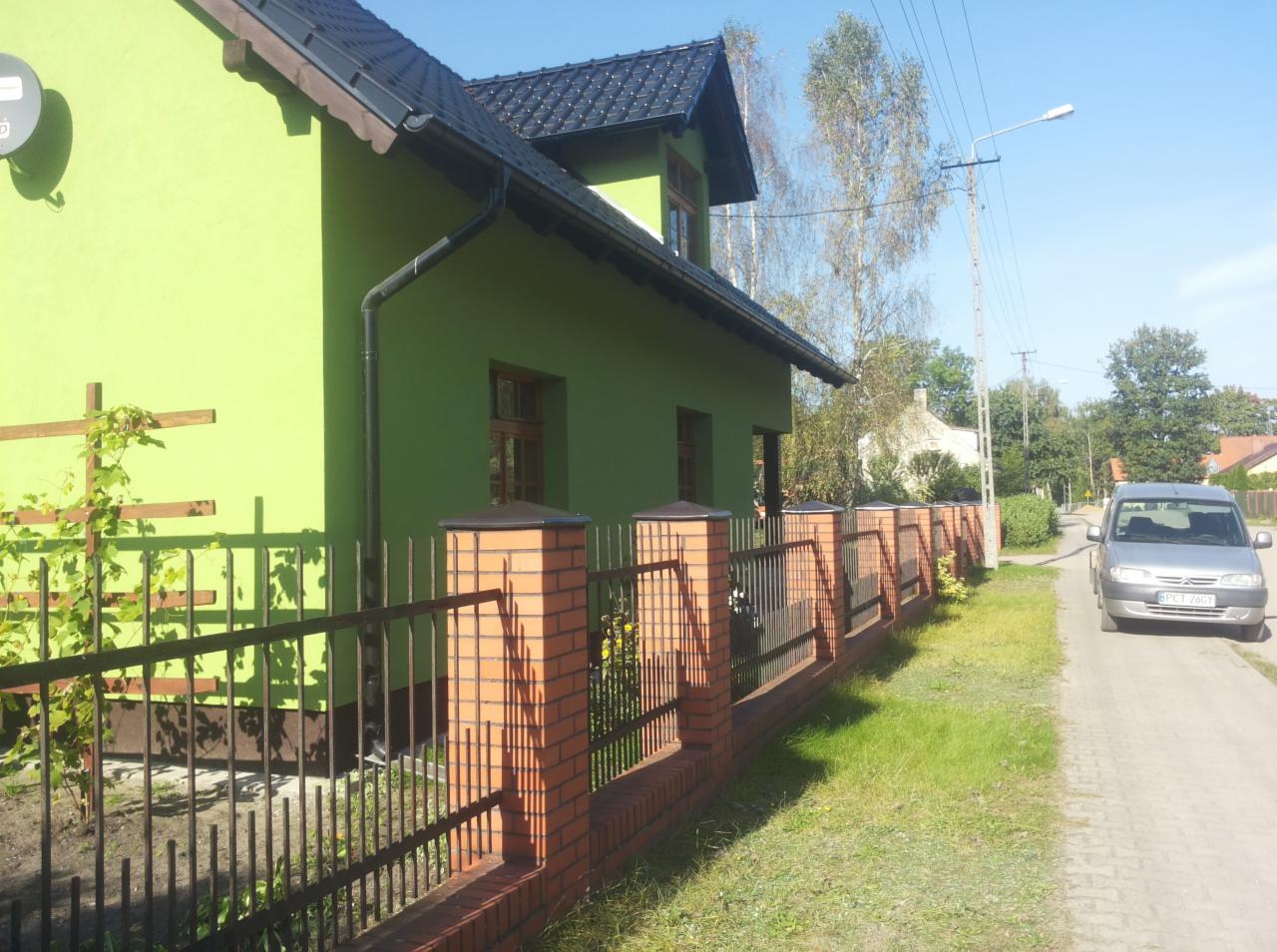 Zielony dom murowany z ciemną dachówką, widoczny zza ceglanego ogrodzenia z metalowymi przęsłami, na tle błękitnego nieba i ulicy z zaparkowanym srebrnym samochodem.