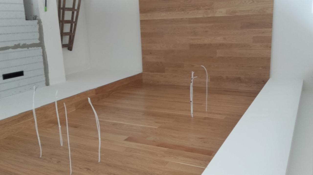 Jasnobrązowa drewniana podłoga ułożona w deski, widoczne wystające kable elektryczne, fragment ściany wykończonej drewnem oraz surowy fragment ściany z instalacją elektryczną w tle.