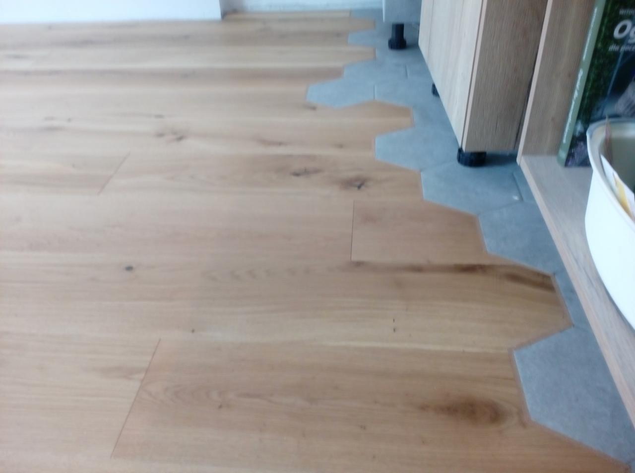 Jasna drewniana podłoga ułożona z desek, przechodząca w szare heksagonalne płytki pod szafkami kuchennymi.