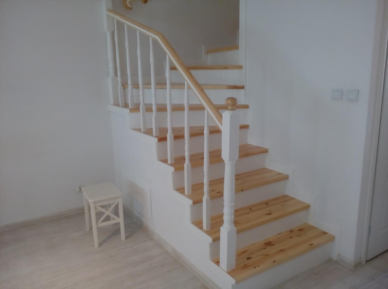 Jasne drewniane schody z białymi balustradami w minimalistycznym wnętrzu, widoczny mały biały stołek obok schodów.