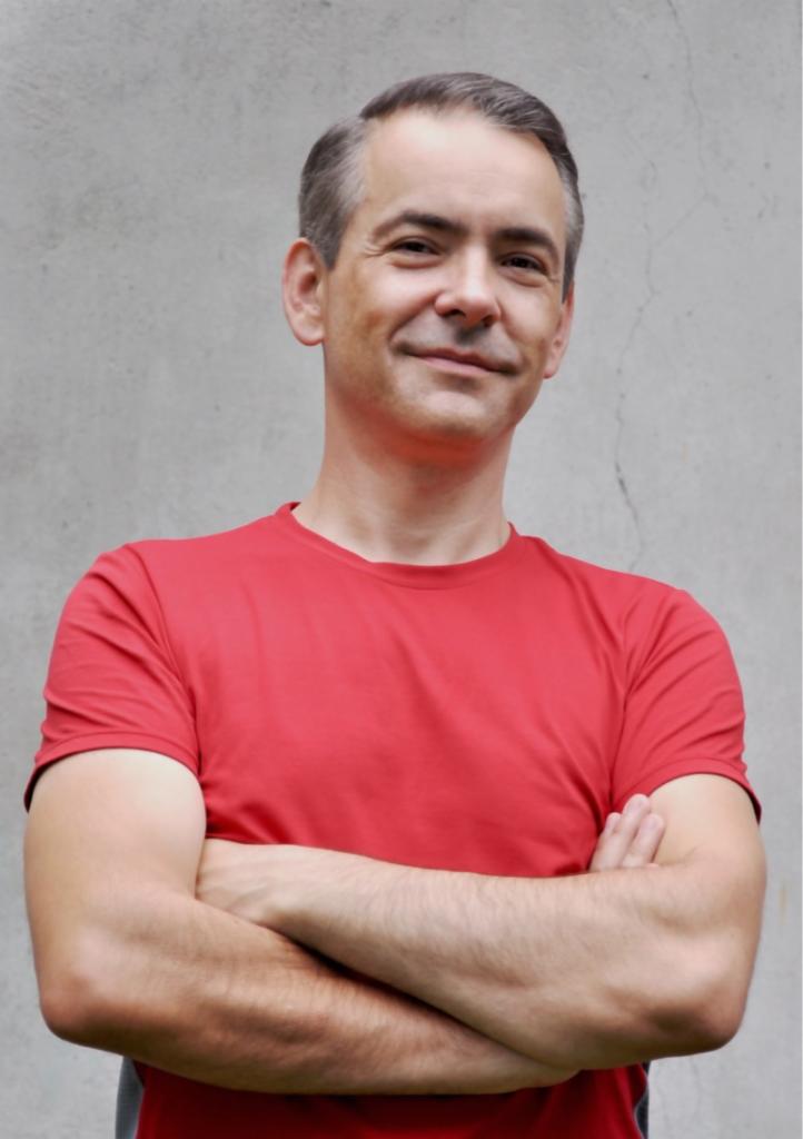 Portret uśmiechniętego mężczyzny w czerwonej koszulce z założonymi rękami na tle szarej ściany.
