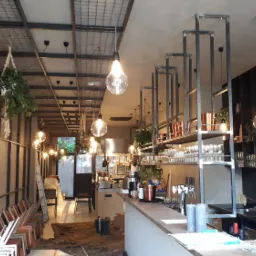 Wnętrze baru w trakcie wykańczania: widoczne wiszące lampy, metalowe regały z naczyniami, bar, plan architektoniczny na ścianie, drabina i krzesła w tle.