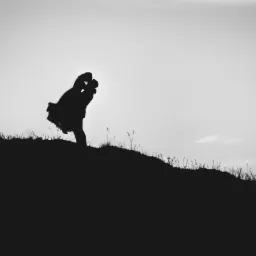 Czarno-biała sylwetka pary całującej się na szczycie wzgórza porośniętego trawą, na tle jasnego nieba.