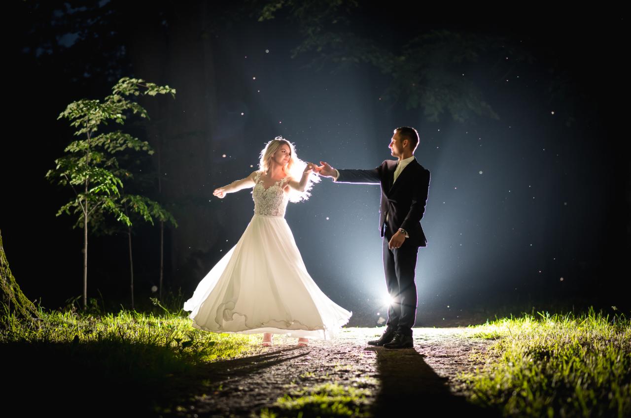 Para młoda tańczy w nocnej scenerii leśnej, oświetlona punktowym światłem, tworzącym magiczną atmosferę z widocznymi drobinkami w powietrzu.