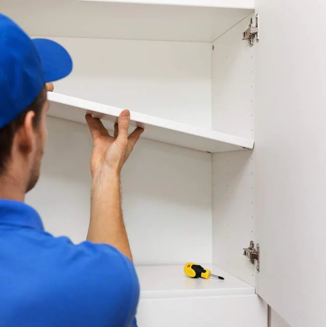 Montaż białej półki w szafce przez montera w niebieskiej czapce, widoczny żółto-czarny śrubokręt na dolnej półce.