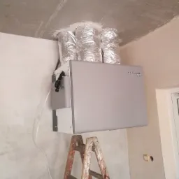 Szara jednostka rekuperacji DEFRO AR zamontowana pod surowym betonowym sufitem, widoczne trzy srebrne, izolowane przewody wentylacyjne oraz drabina malarska.