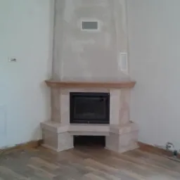 Narożny, obudowany jasnym kamieniem z czarnym wkładem, z drewnianą półką, kominek z pionowym, surowym wykończeniem ściany kominowej z otworami wentylacyjnymi, na tle drewnianej podłogi.