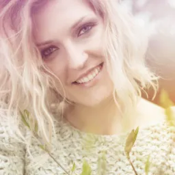 Uśmiechnięta blondynka z delikatnym makijażem i kręconymi włosami, ubrana w gruby, wełniany sweter, pozuje na tle rozmytej zieleni i różu.