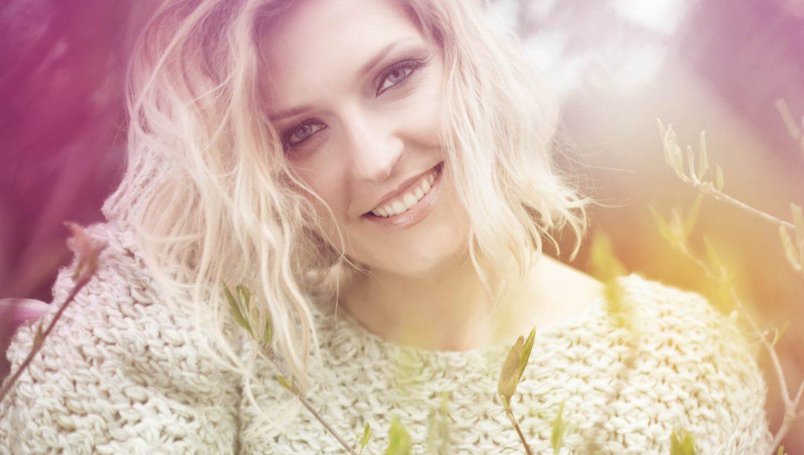 Uśmiechnięta blondynka z delikatnym makijażem i kręconymi włosami, ubrana w gruby, wełniany sweter, pozuje na tle rozmytej zieleni i różu.