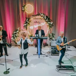 Zespół muzyczny 'Alibi' z Lubartowa podczas występu na scenie z dekoracją kwiatową i podświetlanym napisem. Wokalistka z mikrofonem, klawiszowiec, gitarzysta i saksofonista.