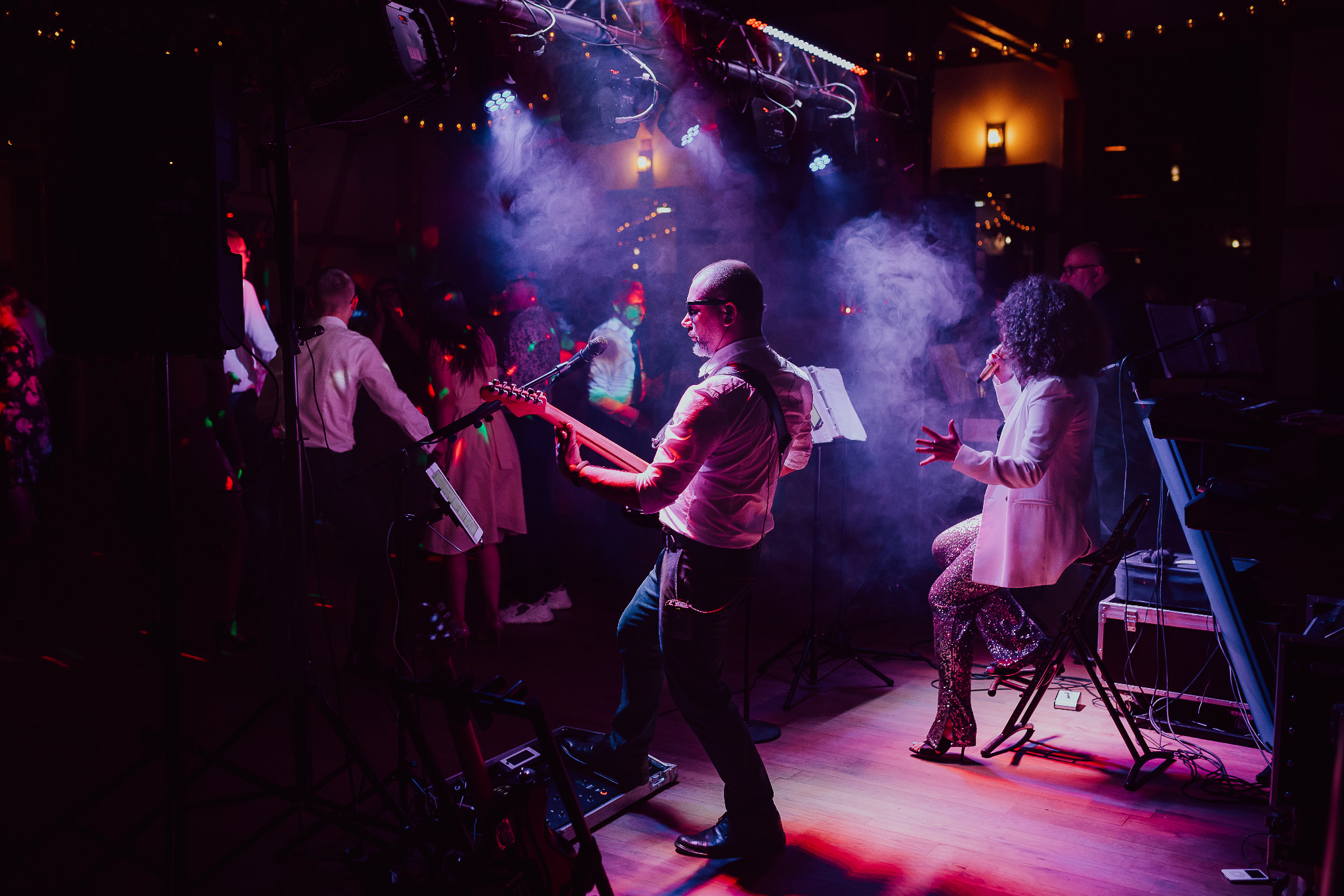 Zespół muzyczny grający na żywo podczas imprezy, gitarzysta w okularach przeciwsłonecznych i w białej koszuli, wokalistka w cekinowych spodniach, dynamiczne oświetlenie sceniczne, w tle tańczący tłum.