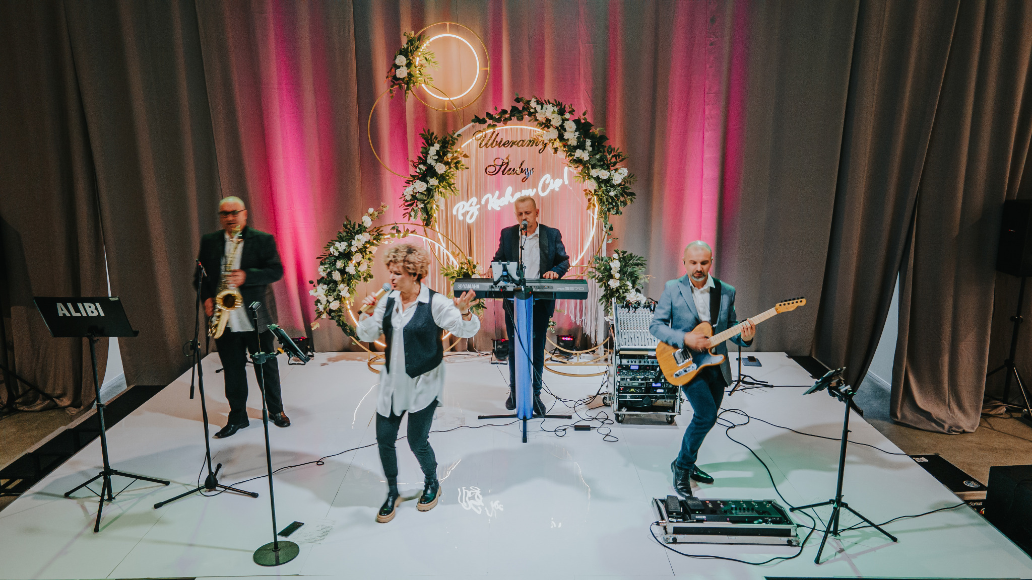 Zespół muzyczny 'Alibi' z Lubartowa podczas występu na scenie z dekoracją kwiatową i podświetlanym napisem. Wokalistka z mikrofonem, klawiszowiec, gitarzysta i saksofonista.