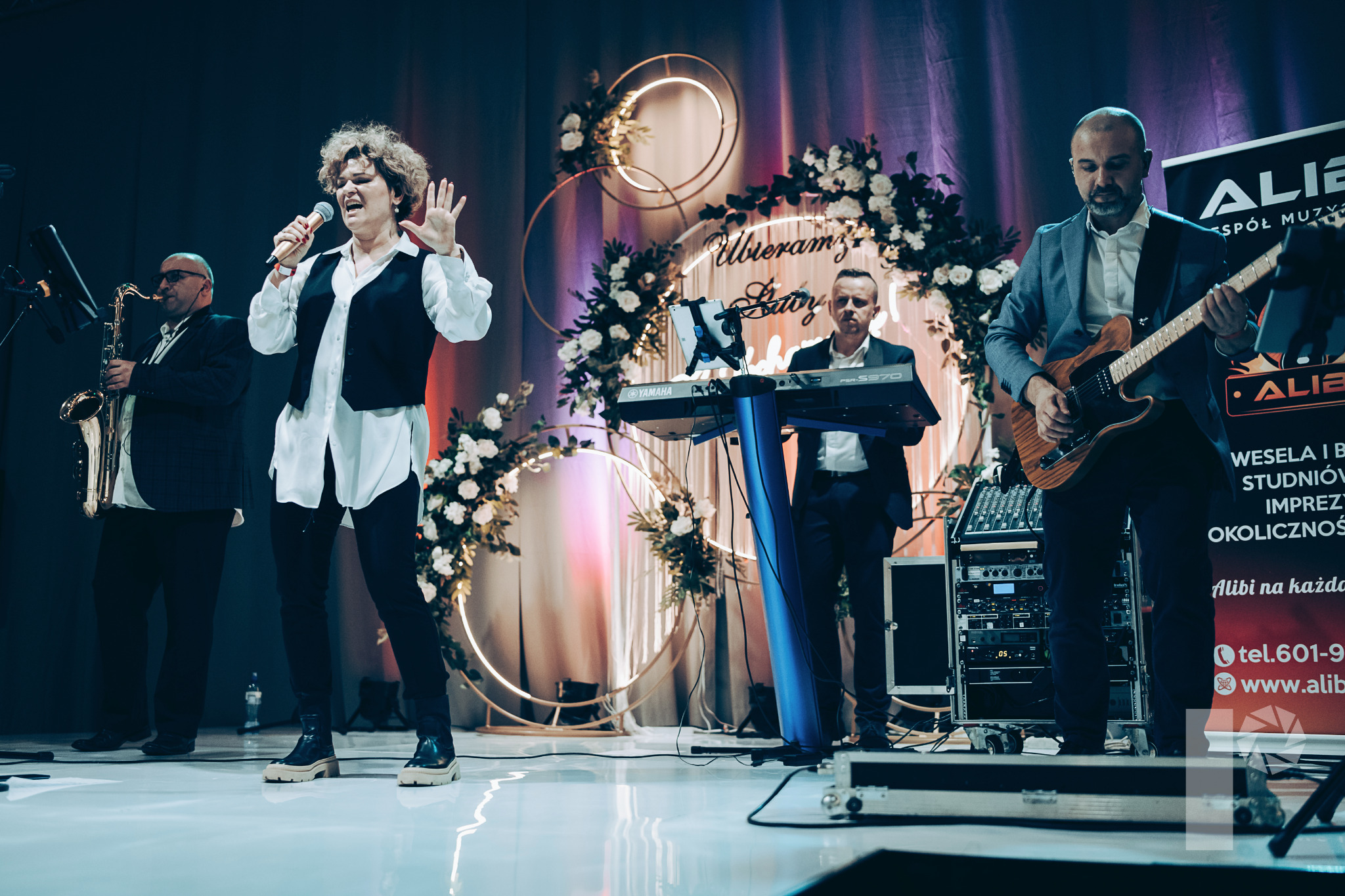 Zespół muzyczny na scenie podczas występu: wokalistka z mikrofonem, saksofonista, klawiszowiec grający na Yamaha PSR-S970 i gitarzysta, dekoracja z kwiatów i obręczy w tle.