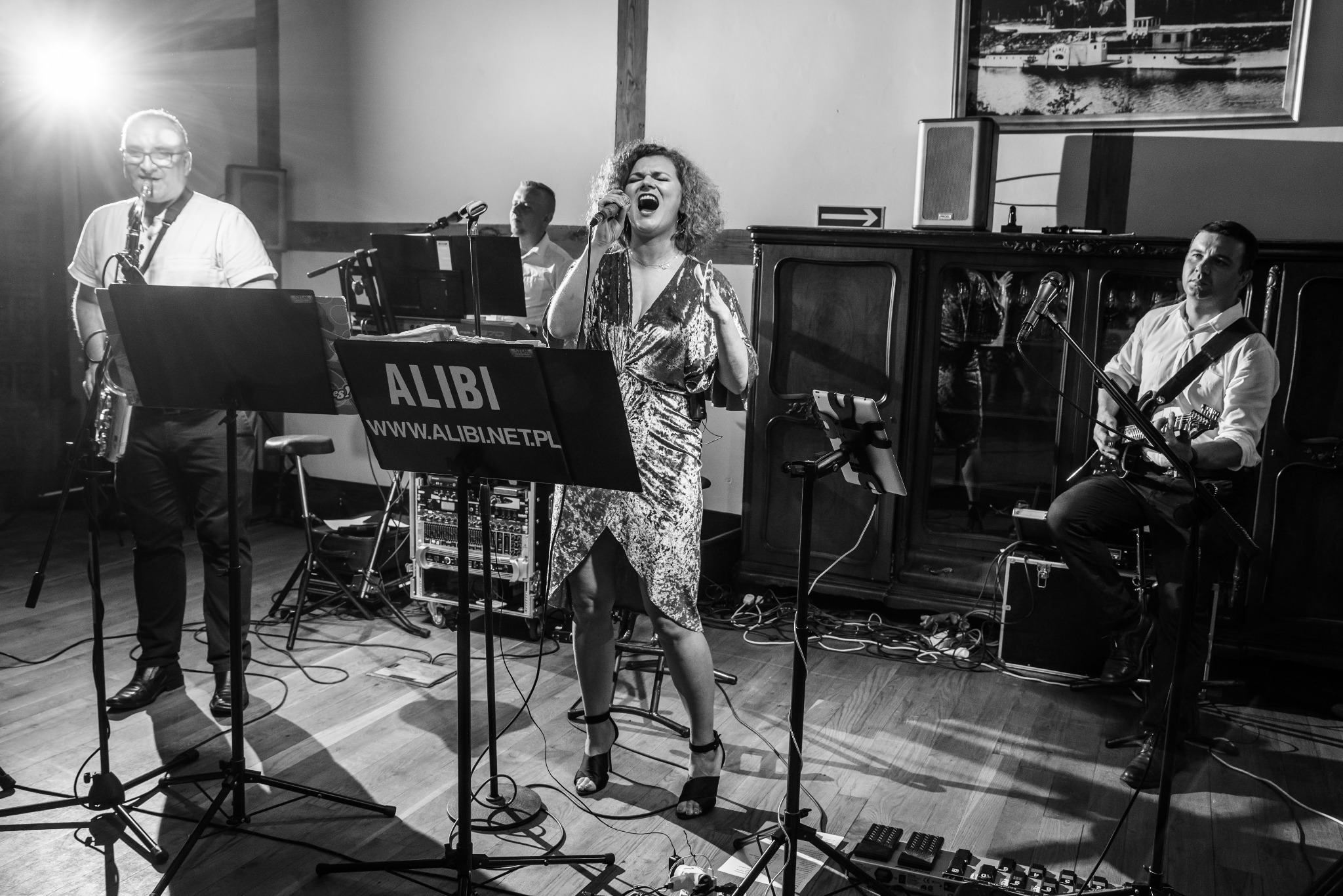 Czarno-białe zdjęcie zespołu muzycznego podczas występu: wokalistka śpiewa z ekspresją, saksofonista z lewej strony gra, gitarzysta siedzi po prawej, za nimi klawiszowiec. Widoczny baner z nazwą...