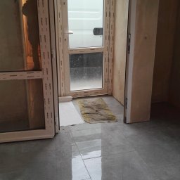 MIREX Mirosław Koziana - Nowe, szare płytki podłogowe z połyskiem, widoczne otwarte, białe drzwi balkonowe i fragment ściany w trakcie prac wykończeniowych.