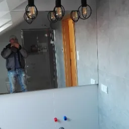 Nowoczesna łazienka w szaro-białej kolorystyce z industrialną lampą wiszącą z żarówkami Edisona, widoczne przyłącza wodne i odbicie osoby robiącej zdjęcie w lustrze.