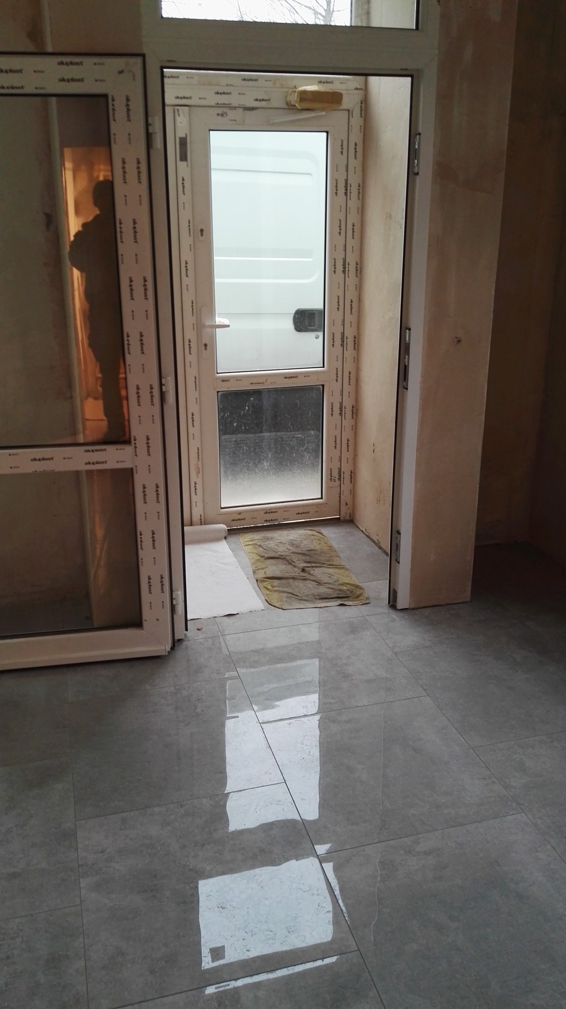 Nowe, szare płytki podłogowe z połyskiem, widoczne otwarte, białe drzwi balkonowe i fragment ściany w trakcie prac wykończeniowych.