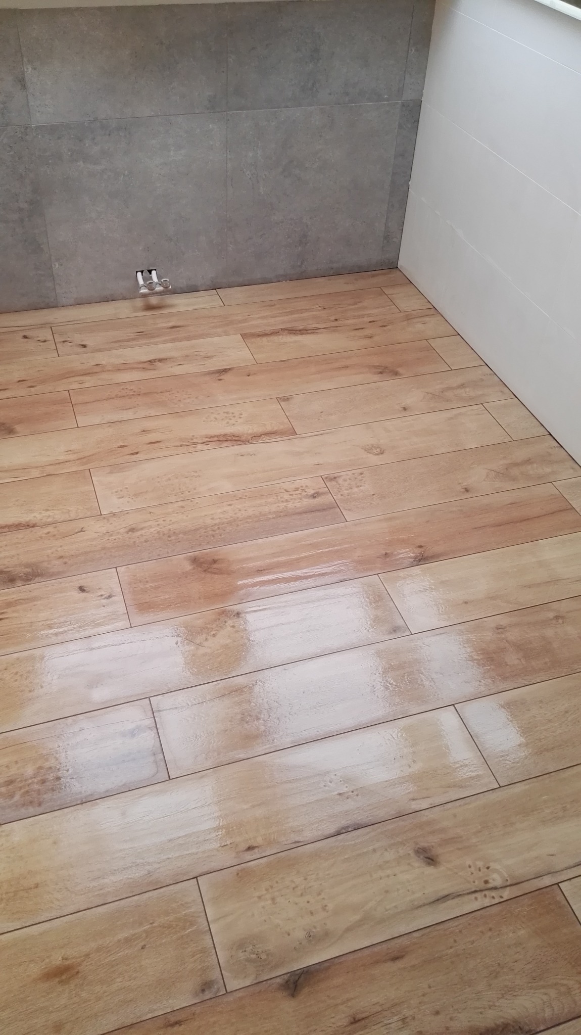 Ułożona podłoga z płytek imitujących drewno w łazience, szare płytki ścienne i białe panele ścienne, widoczne rury instalacji wodnej.