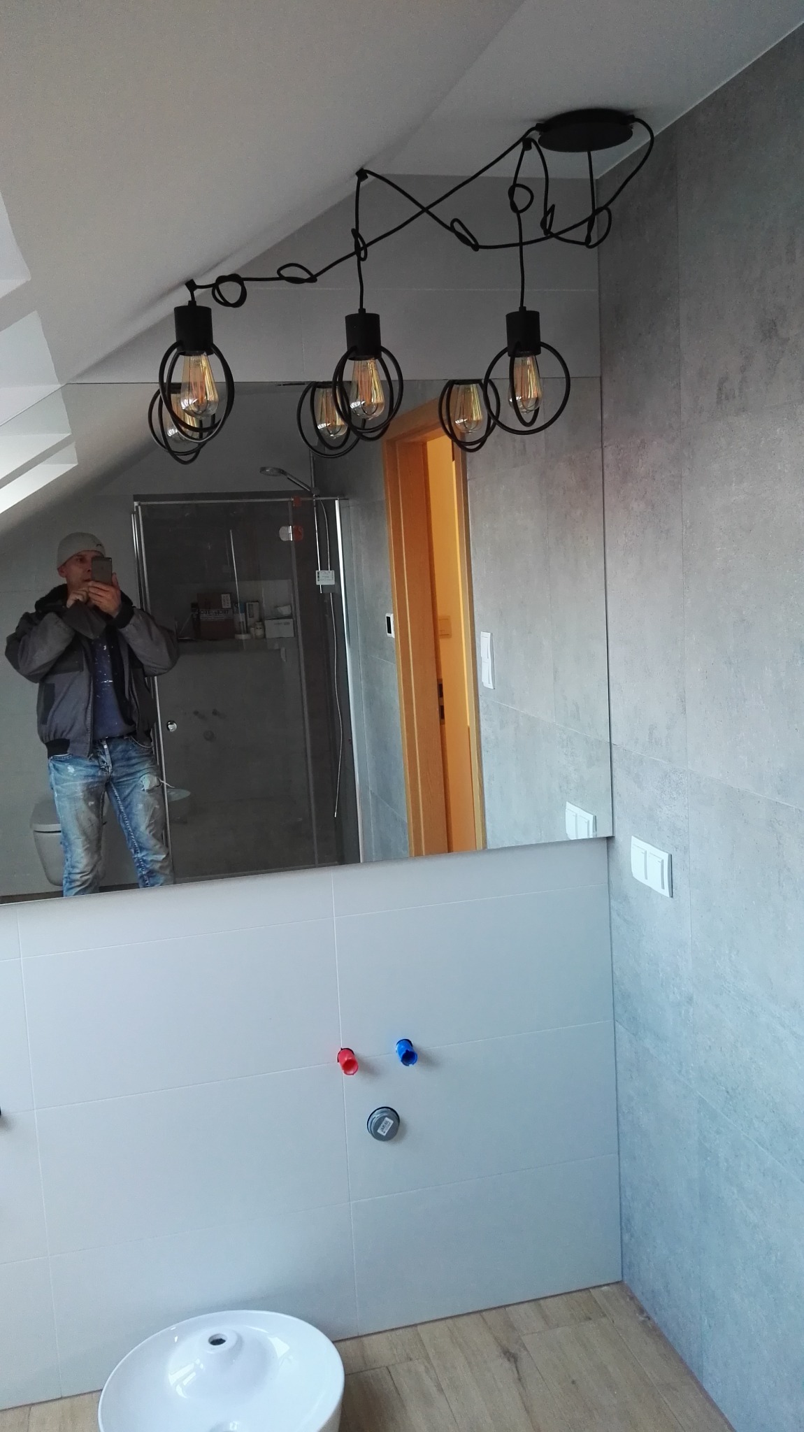 Nowoczesna łazienka w szaro-białej kolorystyce z industrialną lampą wiszącą z żarówkami Edisona, widoczne przyłącza wodne i odbicie osoby robiącej zdjęcie w lustrze.