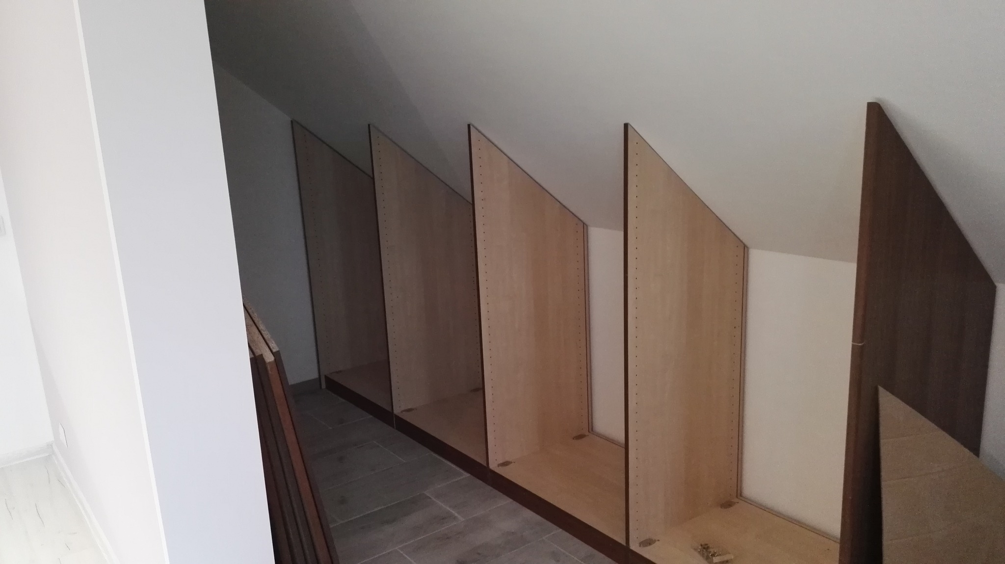 Szkielet zabudowy wnęki ze skośnym sufitem, widoczne płyty z otworami na kołki i brązowe ramy, szare płytki podłogowe.