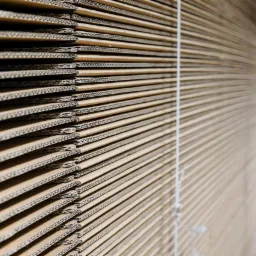 Stos płaskich, złożonych kartonów z widoczną strukturą tektury falistej, spiętych sznurkiem, na tle magazynu.