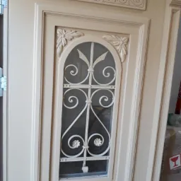 Beżowe drzwi wewnętrzne z ozdobnym, białym, metalowym wzorem w szybie, widoczne detale dekoracyjne na ramie i górnej listwie.