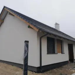 Nowo otynkowany, parterowy dom murowany z czarną dach&oacute;wką i orynnowaniem, okna z roletami zewnętrznymi, w tle pochmurne niebo.