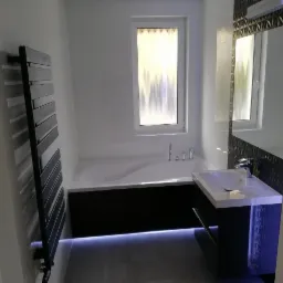 Nowoczesna łazienka z białą wanną obudowaną ciemnobrązową mozaiką, białą umywalką z szafką i podświetleniem LED u dołu, grzejnikiem drabinkowym oraz lustrem z geometrycznym wzorem.