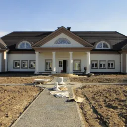 Elegancki, parterowy dom z kolumnami i symetrycznym układem okien, widok z przodu, z częściowo ułożoną kostką brukową na podjeździe i przygotowanym terenem pod trawnik.