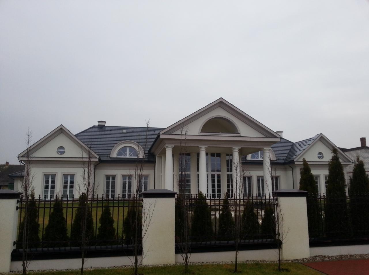 Elegancki, biały dom z kolumnami i czarną, kutą bramą, otoczony młodymi drzewkami iglastymi i trawnikiem, sfotografowany w pochmurny dzień.