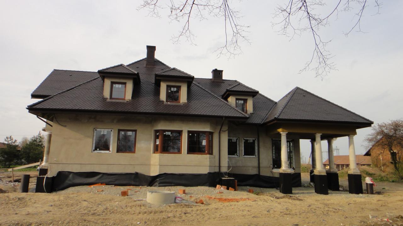 Niedokończony dom murowany z brązowym dachem, widoczne okna z brązowymi ramami i kolumnowy ganek, teren wokół domu w trakcie przygotowania, ziemia z cegłami.