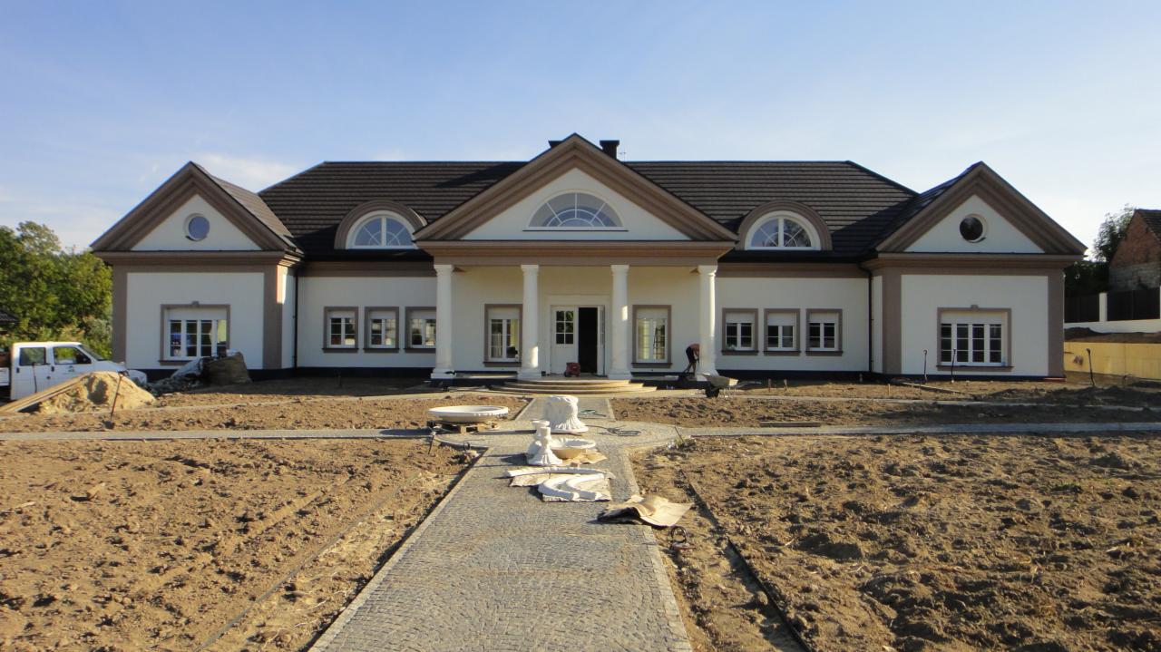 Elegancki, parterowy dom z kolumnami i symetrycznym układem okien, widok z przodu, z częściowo ułożoną kostką brukową na podjeździe i przygotowanym terenem pod trawnik.