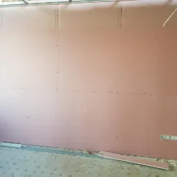 Różowa ściana z płyt gipsowo-kartonowych na metalowym stelażu, widoczne łączenia płyt i otwory na gniazdka elektryczne, fragment konstrukcji sufitu podwieszanego.