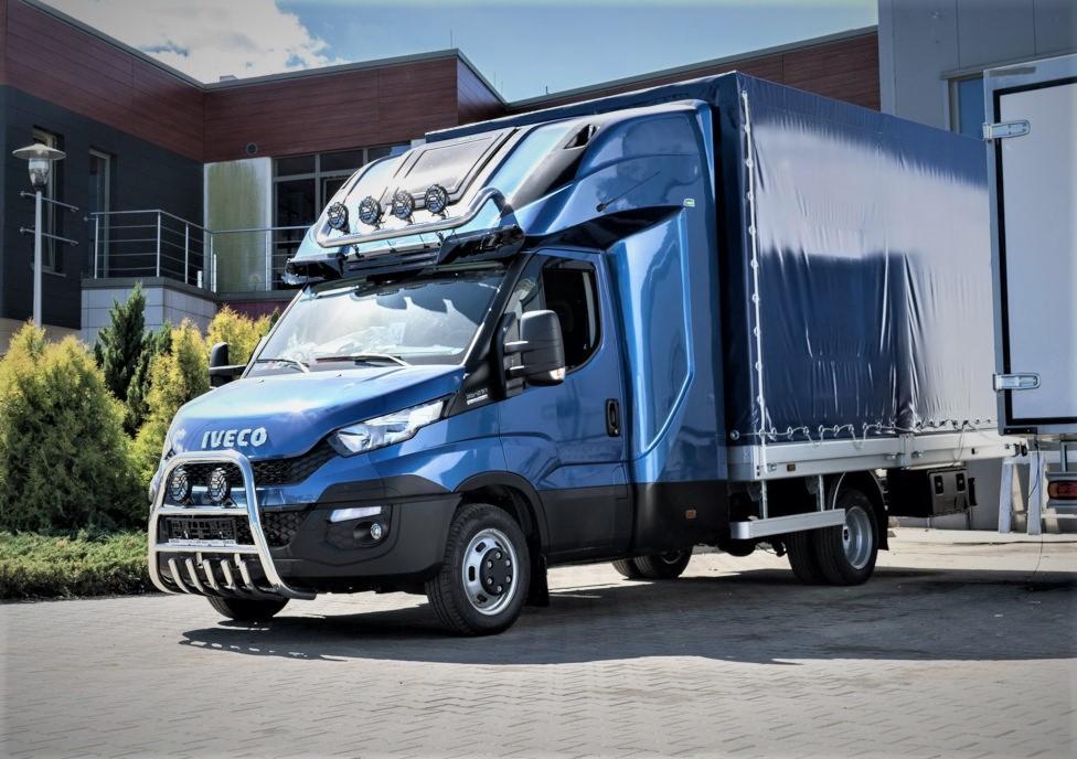 Niebieski samochód dostawczy Iveco z plandeką, dodatkowym oświetleniem i orurowaniem ochronnym na tle nowoczesnego budynku.