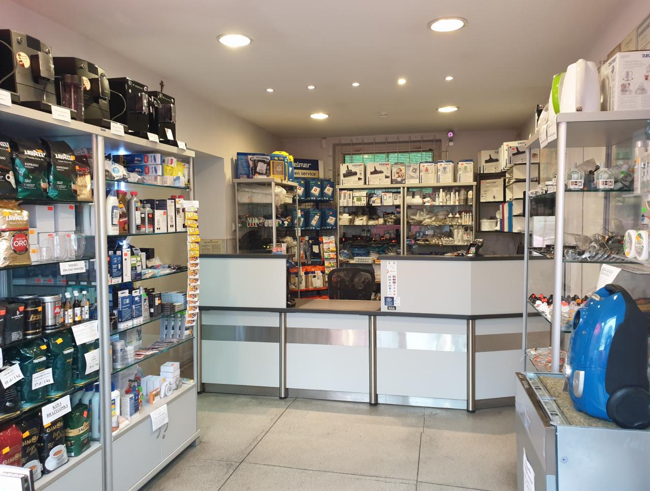 Wnętrze sklepu z asortymentem AGD, ekspresami do kawy i akcesoriami, widok na ladę i regały z produktami.