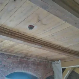 Drewniany sufit z belkami konstrukcyjnymi i ozdobną listwą, częściowo pomalowany, fragment muru z cegły z łukowym przejściem widoczny w dolnej części kadru.