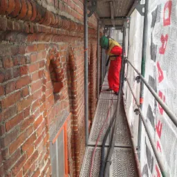 Pracownik w czerwonym kombinezonie i zielonym hełmie na rusztowaniu czyści ceglaną elewację zabytkowej kamienicy. Widoczne elementy dekoracyjne z cegły i zabezpieczająca siatka z nadrukiem.