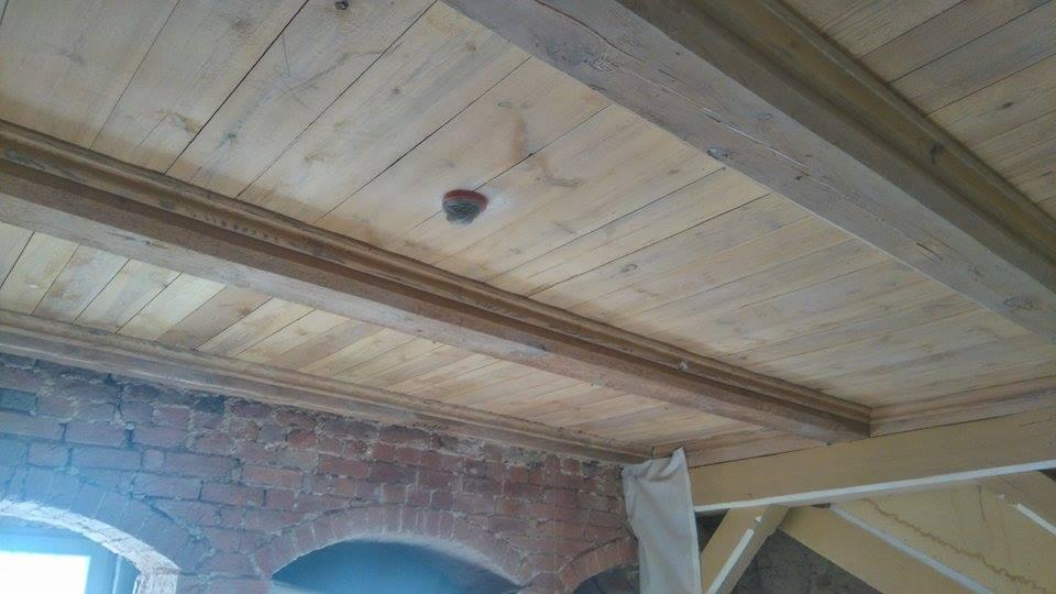 Drewniany sufit z belkami konstrukcyjnymi i ozdobną listwą, częściowo pomalowany, fragment muru z cegły z łukowym przejściem widoczny w dolnej części kadru.