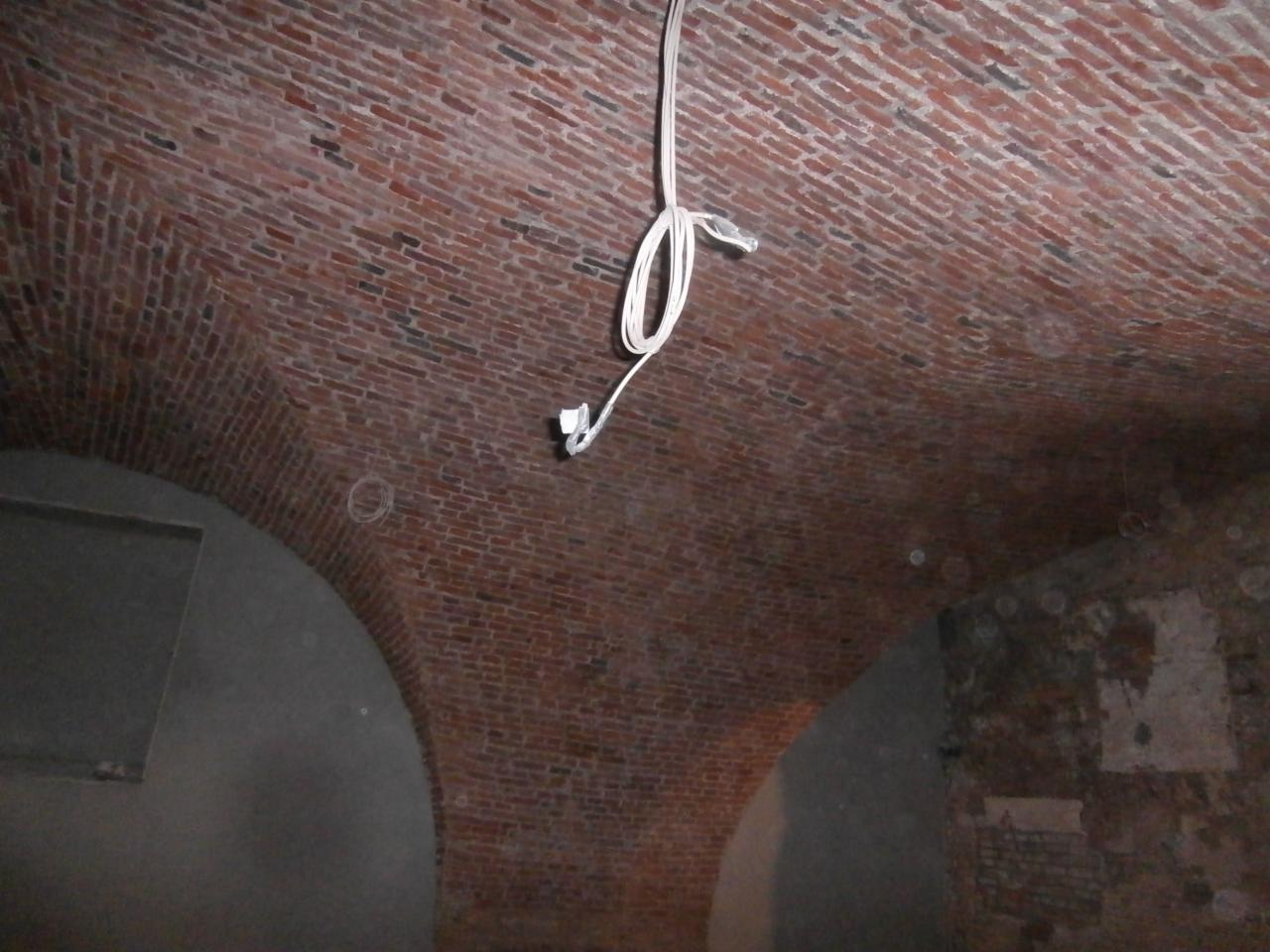 Ceglany, łukowy strop piwnicy w trakcie renowacji z wiszącymi kablami elektrycznymi. Widoczne fragmenty otynkowanych ścian i ślady wcześniejszych prac.