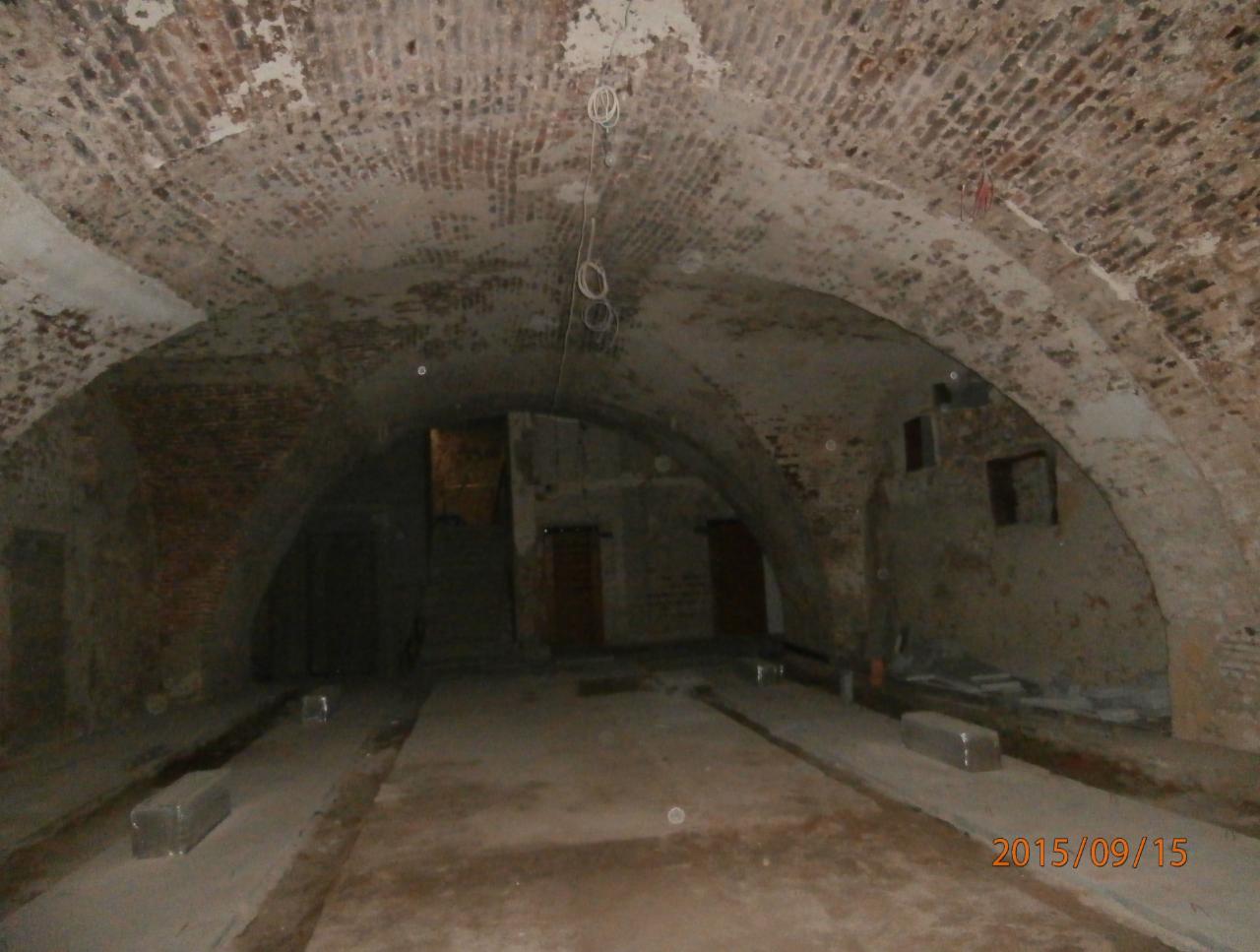 Ciemne, sklepione piwnice z ceglanym sufitem, widoczne ślady wilgoci i zniszczeń, betonowa podłoga z metalowymi elementami, słabe oświetlenie.