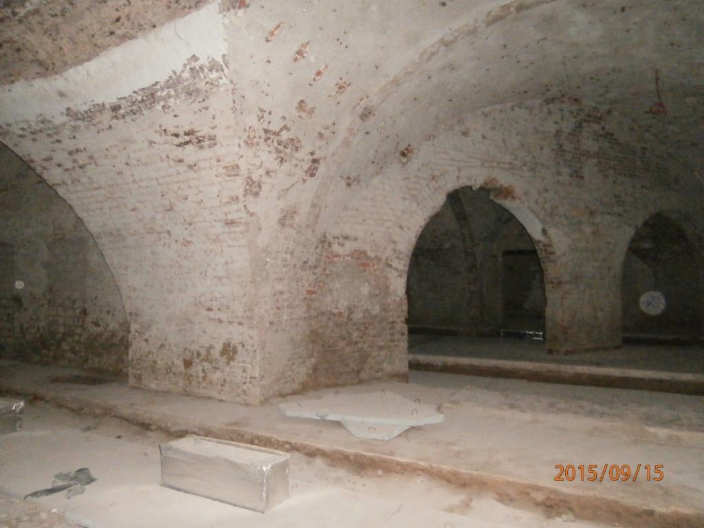 Ceglane sklepienie łukowe w piwnicy, widoczne ślady wilgoci i napraw, posadzka betonowa, w tle przejścia między pomieszczeniami.