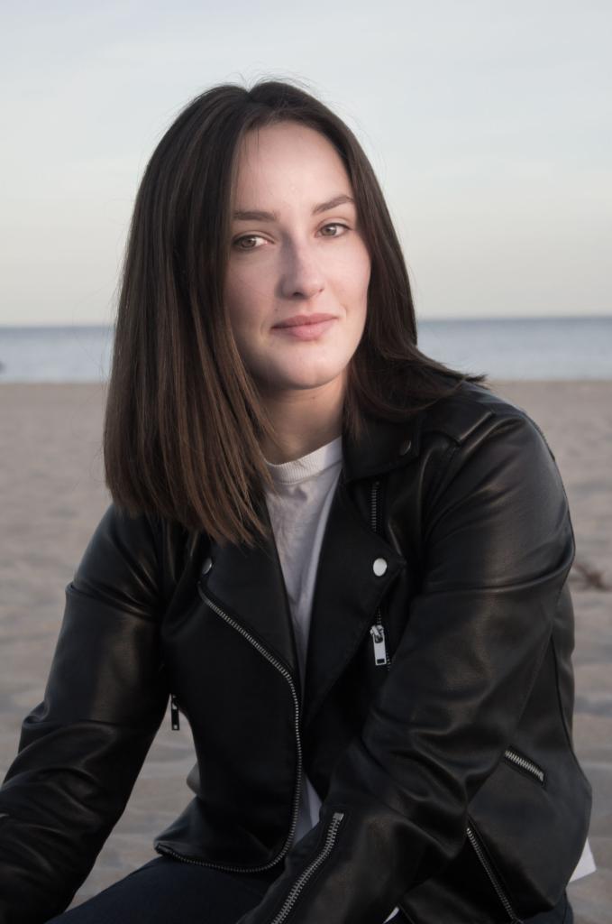 Portret kobiety w czarnej skórzanej kurtce na plaży, z widocznym piaskiem i morzem w tle, delikatne oświetlenie.