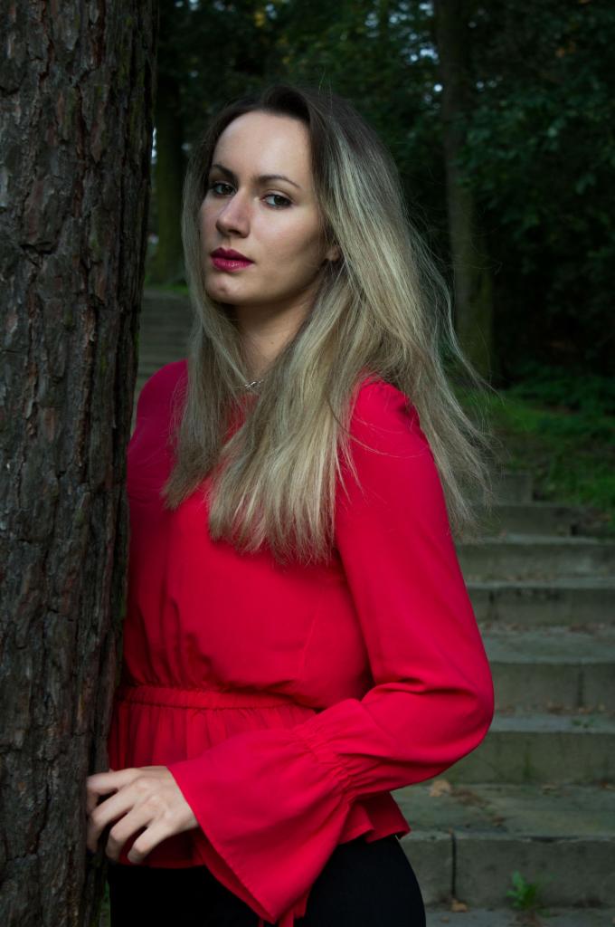 Portret kobiety w czerwonej bluzce, opierającej się o korę drzewa na tle schodów w parku.