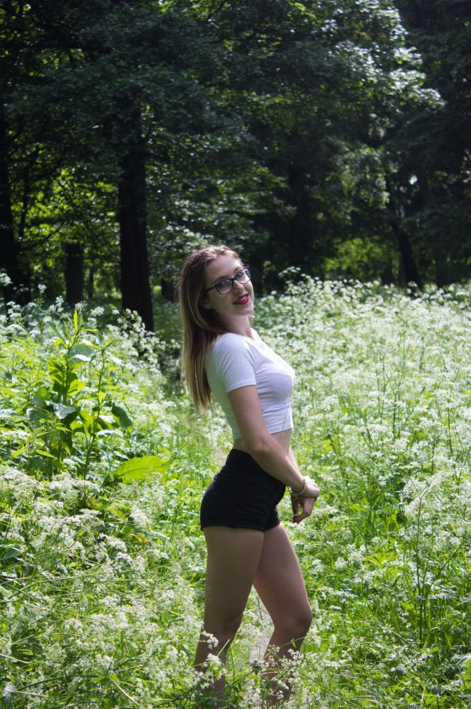 Uśmiechnięta młoda kobieta w okularach, białym topie i czarnych szortach pozuje w plenerze, otoczona kwitnącymi białymi kwiatami i zielenią drzew.