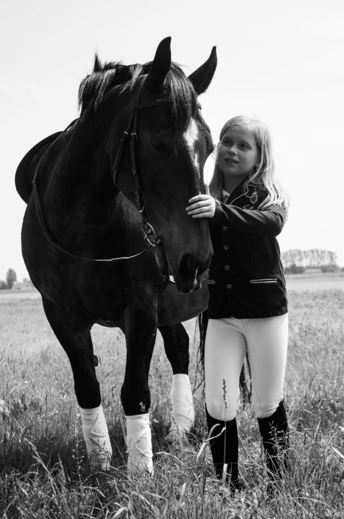 Czarno-białe zdjęcie dziewczynki w stroju jeździeckim, delikatnie dotykającej pyska ciemnego konia na łące.