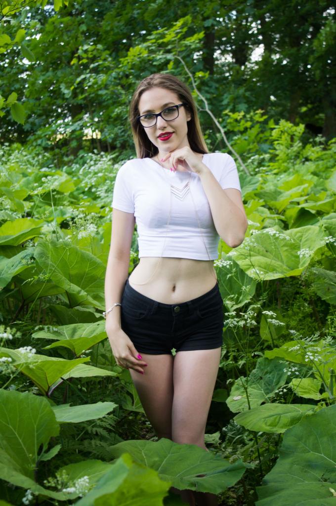 Kobieta w okularach, białym topie i czarnych szortach pozuje w plenerze wśród zieleni.
