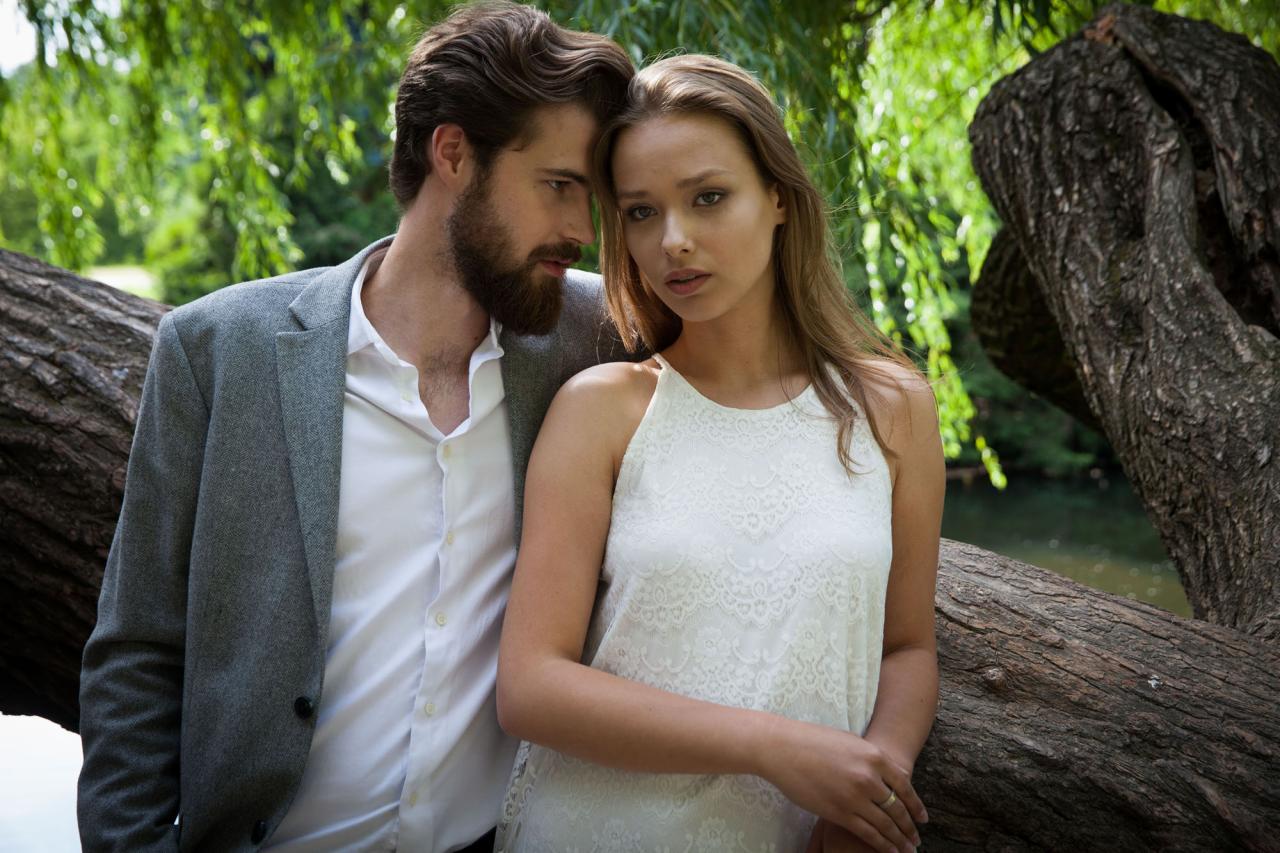 Para zakochanych, mężczyzna w szarej marynarce i kobieta w koronkowej sukience, opierają się o stare drzewo nad wodą, w tle zieleń parku.