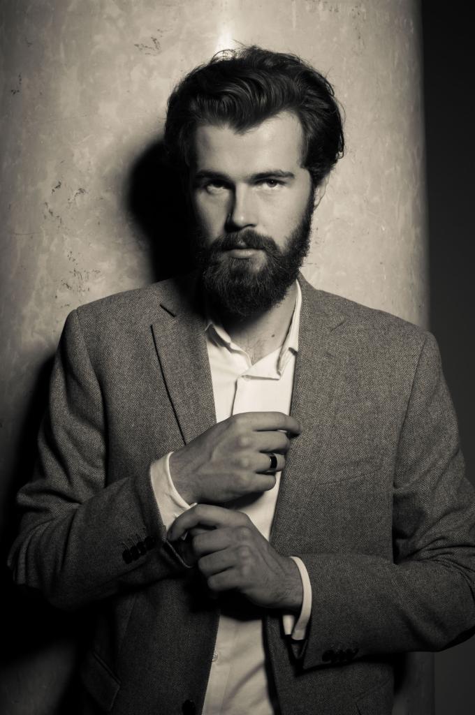 Portret brodatego mężczyzny w tweedowej marynarce, poprawiającego mankiet koszuli, stojącego przy marmurowej kolumnie w czarno-białej tonacji.